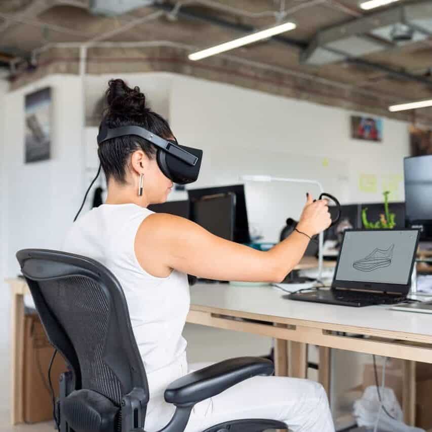 Eine Frau sitzt vor einem Laptop und trägt eine VR-Brille. Sie bearbeitet 3D-Rundgänge. 